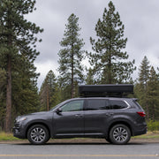 Closed TrailNest Panorama rooftop tent mounted on a gray SUV beside a forest road