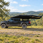 Vehicle-mounted fan awning on a black pickup at an outdoor campsite - illustrative scene based on supplier reference photos